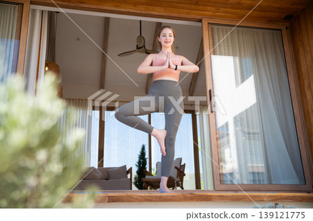 Fit woman practicing balancing yoga pose on modern house balcony Fit woman practicing balancing yoga pose on modern house balcony 139121775