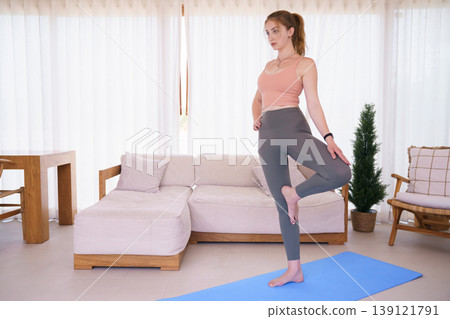 Young woman in sports bra practicing tree pose yoga at home Young woman in sports bra practicing tree pose yoga at home 139121791