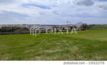 Panoramic view of open green Maltese landscape with rolling fields in foreground and modern construction buildings breaking natural horizon under cloudy blue sky 139122776