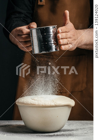 Sifting flour over risen dough in bowl with baker hands on dark background Sifting flour over risen dough in bowl with baker hands on dark background 139124020