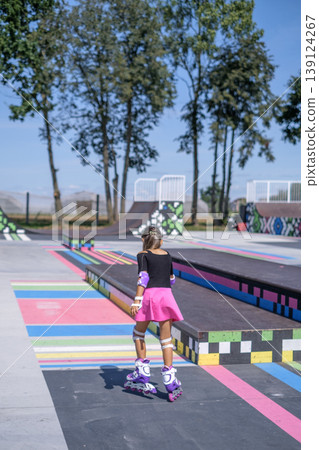 The girl confidently and professionally roller skates on the playground of the skate park. Rear view. The girl confidently and professionally roller skates on the playground of the skate park. Rear view. 139124267