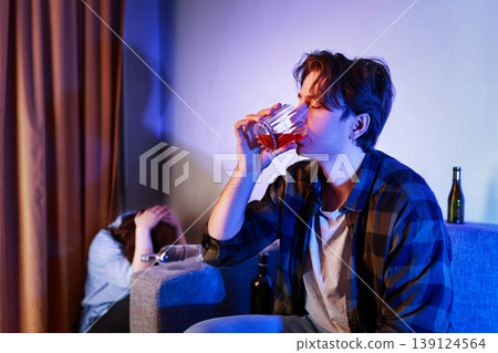 Drunk man drinks alcohol from a glass while his crying wife hides behind the sofa. Concept of alcoholism, addiction, domestic violence, and a ruined family life. Drunk man drinks alcohol from a glass while his crying wife hides behind the sofa. Concept of alcoholism, addiction, domestic violence, and a ruined family life. 139124564