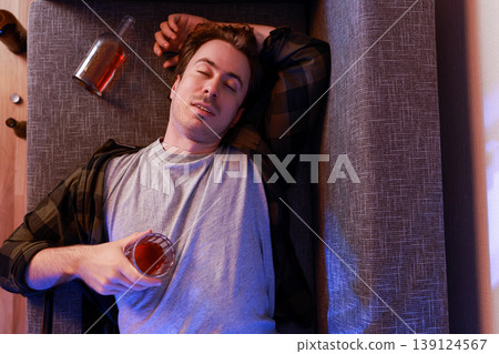 Top view of an unconscious drunk man asleep on his back on a sofa, holding a glass of alcohol. Empty bottles around him. Concept of severe addiction, divorce, or unemployment. 139124567