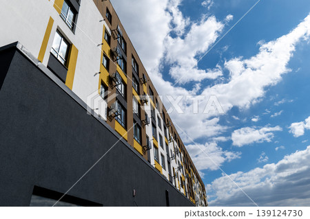 Sunlight filters through fluffy clouds as a stylish building stands tall, revealing artful designs in its facade and a vibrant city atmosphere 139124730