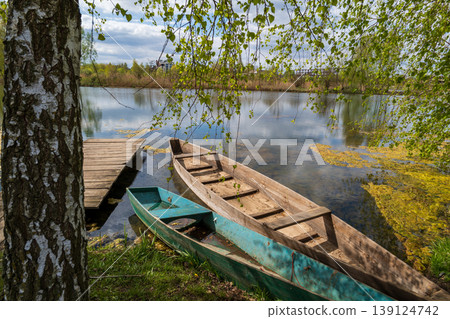 Two wooden boats are nestled by the calm lake, surrounded by lush greenery and reflecting clouds in the serene water Two wooden boats are nestled by the calm lake, surrounded by lush greenery and reflecting clouds in the serene water 139124742