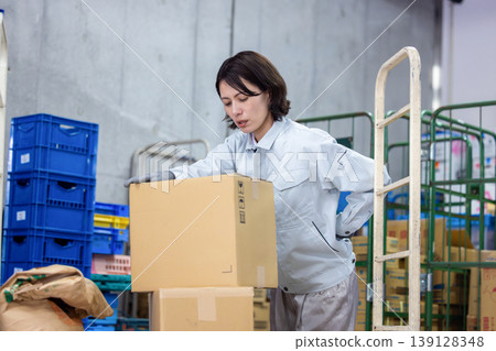 A woman carrying a cardboard cart in a warehouse. 139128348