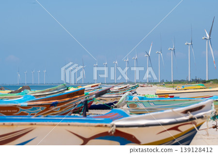 Colorful fishing boats and offshore wind turbines under a clear blue sky. Coastal wind farm on Talaimannar beach, Sri Lanka. Colorful fishing boats and offshore wind turbines under a clear blue sky. Coastal wind farm on Talaimannar beach, Sri Lanka. 139128412