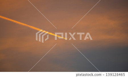 Airplane contrail streaking across a dramatic orange sunset sky with soft clouds and warm golden light, minimalist atmospheric background Airplane contrail streaking across a dramatic orange sunset sky with soft clouds and warm golden light, minimalist atmospheric background 139128465