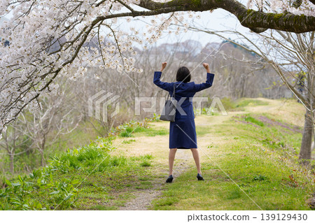 一位身穿西裝的女子背對著鏡頭，在櫻花樹下擺出勝利的姿勢。 139129430