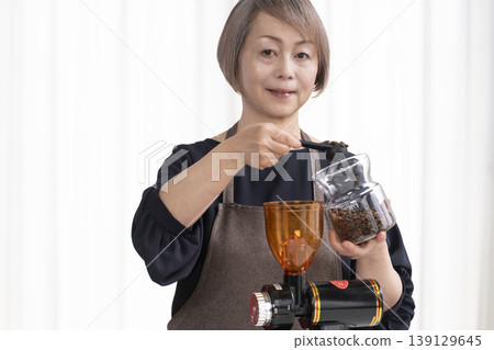 A senior woman grinds coffee beans and drinks coffee in her living room. A senior woman grinds coffee beans and drinks coffee in her living room. 139129645