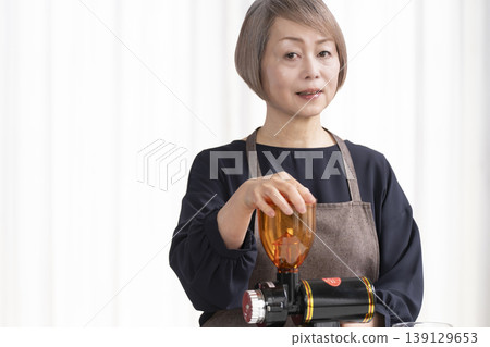 A senior woman grinds coffee beans and drinks coffee in her living room. A senior woman grinds coffee beans and drinks coffee in her living room. 139129653