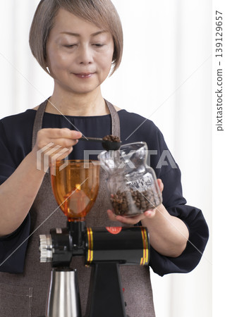 A senior woman grinds coffee beans and drinks coffee in her living room. A senior woman grinds coffee beans and drinks coffee in her living room. 139129657