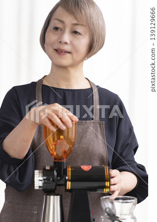 A senior woman grinds coffee beans and drinks coffee in her living room. A senior woman grinds coffee beans and drinks coffee in her living room. 139129666