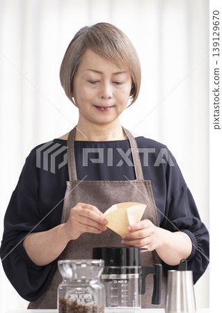A senior woman grinds coffee beans and drinks coffee in her living room. A senior woman grinds coffee beans and drinks coffee in her living room. 139129670