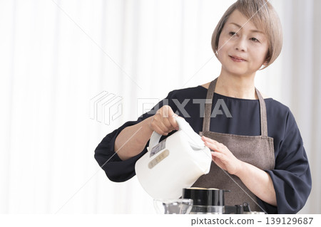 A senior woman grinds coffee beans and drinks coffee in her living room. A senior woman grinds coffee beans and drinks coffee in her living room. 139129687