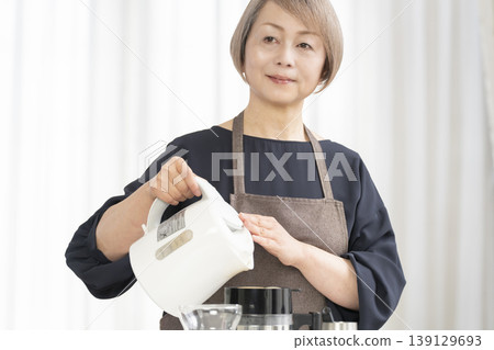 A senior woman grinds coffee beans and drinks coffee in her living room. A senior woman grinds coffee beans and drinks coffee in her living room. 139129693