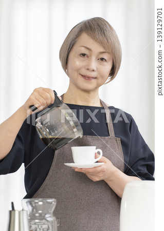 A senior woman grinds coffee beans and drinks coffee in her living room. A senior woman grinds coffee beans and drinks coffee in her living room. 139129701