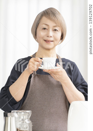 A senior woman grinds coffee beans and drinks coffee in her living room. A senior woman grinds coffee beans and drinks coffee in her living room. 139129702