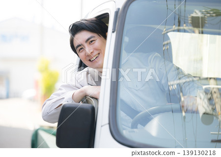 A smiling portrait of a worker leaning out of a car window; outside delivery rounds and on the move, smiling and looking at the camera. A smiling portrait of a worker leaning out of a car window; outside delivery rounds and on the move, smiling and looking at the camera. 139130218