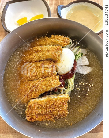 Cold naengmyeon noodles served in a chilled broth with sliced vegetables and egg, paired with crispy golden donkatsu, creating a balance of refreshing and savory flavors in one meal Cold naengmyeon noodles served in a chilled broth with sliced vegetables and egg, paired with crispy golden donkatsu, creating a balance of refreshing and savory flavors in one meal 139134742