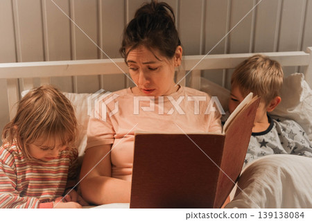 Family bedtime stories. A mother reads a story to her children, wrapped in a blanket. Children listen to soothing stories. A cozy family scene before bed in a softly lit bedroom. Copy space. Banner Family bedtime stories. A mother reads a story to her children, wrapped in a blanket. Children listen to soothing stories. A cozy family scene before bed in a softly lit bedroom. Copy space. Banner 139138084