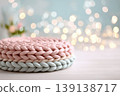 Knitted cushion podium on table, spring blossom bokeh background, pastel tones. Wool pad on surface. Soft pedestal. Cozy stage for promotional imagery. Showcase display case. Front view. Mockup ad 139138717