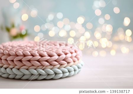 Knitted cushion podium on table, spring blossom bokeh background, pastel tones. Wool pad on surface. Soft pedestal. Cozy stage for promotional imagery. Showcase display case. Front view. Mockup ad 139138717