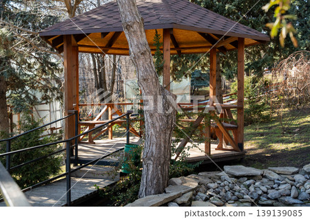 Wooden gazebo with table and benches standing in a park surrounded by trees and greenery with a small bridge and stone landscape in foreground. Outdoor architecture, recreation, leisure Wooden gazebo with table and benches standing in a park surrounded by trees and greenery with a small bridge and stone landscape in foreground. Outdoor architecture, recreation, leisure 139139085