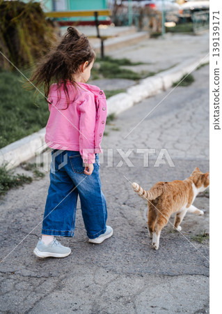 Little girl in pink jacket walking on pavement and following orange cat in residential yard. Concept of childhood curiosity and interaction with animals Little girl in pink jacket walking on pavement and following orange cat in residential yard. Concept of childhood curiosity and interaction with animals 139139171