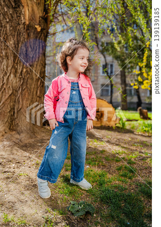Little girl in pink jacket and denim overalls standing near tree in park and smiling while looking aside. Concept of childhood and outdoor lifestyle Little girl in pink jacket and denim overalls standing near tree in park and smiling while looking aside. Concept of childhood and outdoor lifestyle 139139185