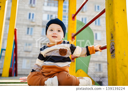 Little boy in hat sitting on playground structure and holding metal bar in sunny yard. Concept of childhood play and outdoor activity Little boy in hat sitting on playground structure and holding metal bar in sunny yard. Concept of childhood play and outdoor activity 139139224