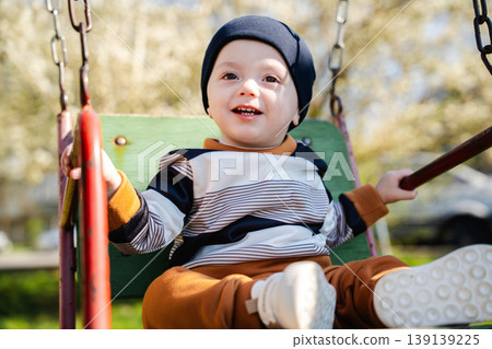 Little boy in hat sitting on swing seat holding bars and smiling in sunny playground environment. Concept of childhood joy and outdoor activity Little boy in hat sitting on swing seat holding bars and smiling in sunny playground environment. Concept of childhood joy and outdoor activity 139139225