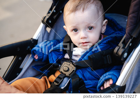 Baby sitting in stroller wearing blue jacket and safety belt looking at camera outdoors. Concept of childcare and safety Baby sitting in stroller wearing blue jacket and safety belt looking at camera outdoors. Concept of childcare and safety 139139227
