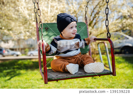 Little boy in hat sitting on swing seat holding bars and looking sideways in sunny playground. Concept of childhood curiosity and outdoor activity 139139228