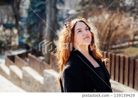 Young caucasian woman with long hair wearing black coat standing outdoors in a park and looking up in sunlight with soft background. Portrait, lifestyle, leisure Young caucasian woman with long hair wearing black coat standing outdoors in a park and looking up in sunlight with soft background. Portrait, lifestyle, leisure 139139429