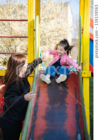 Little girl in pink jacket sliding down playground slide while mother stands nearby and holds her hand. Concept of parenting support and child safety Little girl in pink jacket sliding down playground slide while mother stands nearby and holds her hand. Concept of parenting support and child safety 139139563