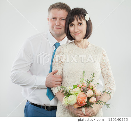 A woman holds a bouquet while hugging a man from behind in a studio. Both smile and look happy. A woman holds a bouquet while hugging a man from behind in a studio. Both smile and look happy. 139140353