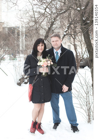 A couple is standing on a snowy street. The woman holds a bouquet of flowers and smiles. A couple is standing on a snowy street. The woman holds a bouquet of flowers and smiles. 139140354