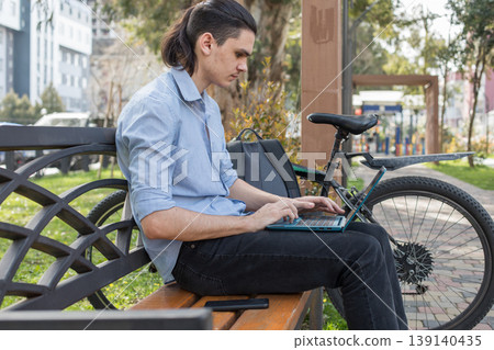A young man sits on a bench in a park during the day. A young man sits on a bench in a park during the day. 139140435