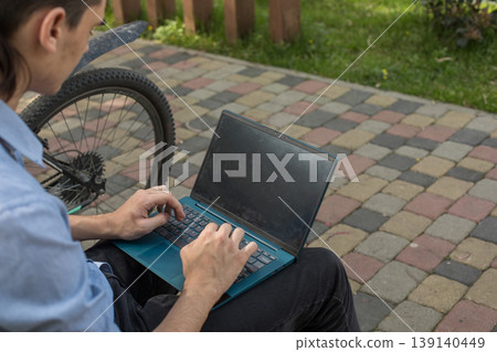 A man sits on a bench in a city park using his laptop. He has a bicycle leaning against the bench A man sits on a bench in a city park using his laptop. He has a bicycle leaning against the bench 139140449