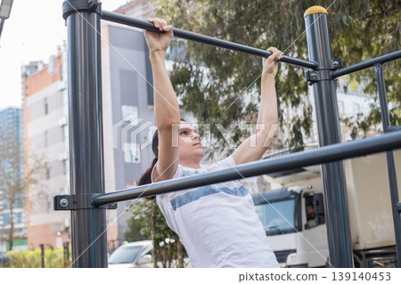 Person does pull-ups at outdoor gym around buildings during daytime 139140453