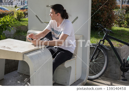 A young man sits on a bench at a bus stop in an urban park. A young man sits on a bench at a bus stop in an urban park. 139140458