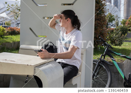 A young man sits on a bench at a bus stop in an urban park. He is focused on his phone A young man sits on a bench at a bus stop in an urban park. He is focused on his phone 139140459