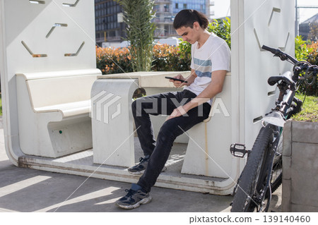 A young man is seated on a bench in an urban space. A young man is seated on a bench in an urban space. 139140460