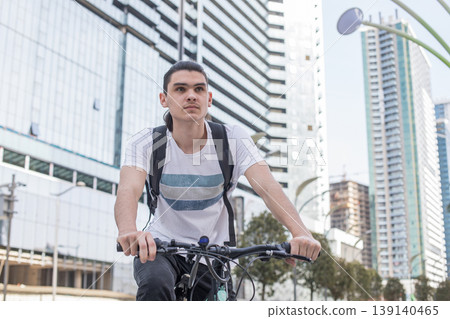 A young man is riding a bicycle in a busy urban area. He wears a simple T-shirt and has a backpack 139140465