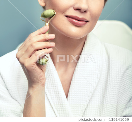 Face jade roller. Cropped face of an adult woman with green jade roller for anti-aging procedures Face jade roller. Cropped face of an adult woman with green jade roller for anti-aging procedures 139141069