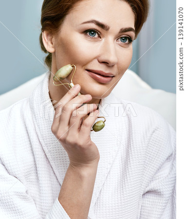 Portrait of a middle-aged woman using jade roller or stone roller for a facial lifting procedure. Close-up Portrait of a middle-aged woman using jade roller or stone roller for a facial lifting procedure. Close-up 139141070