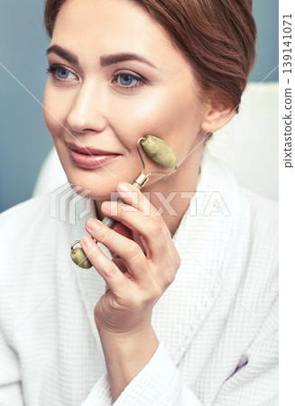 Portrait of a middle-aged woman using jade roller or stone roller for a facial lifting procedure 139141071