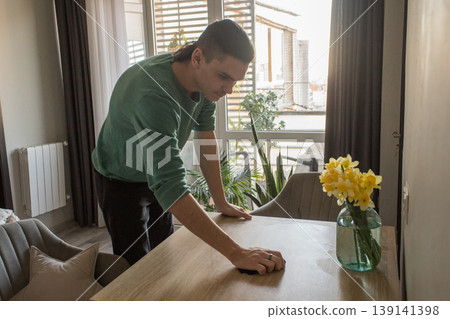 A man is wiping the surface of a wooden table in a well-lit room. A man is wiping the surface of a wooden table in a well-lit room. 139141398