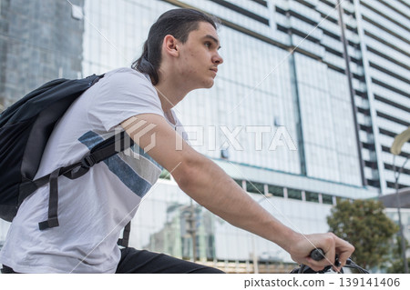 A young man rides a bicycle in a busy city area. Tall buildings surround him as he focuses A young man rides a bicycle in a busy city area. Tall buildings surround him as he focuses 139141406